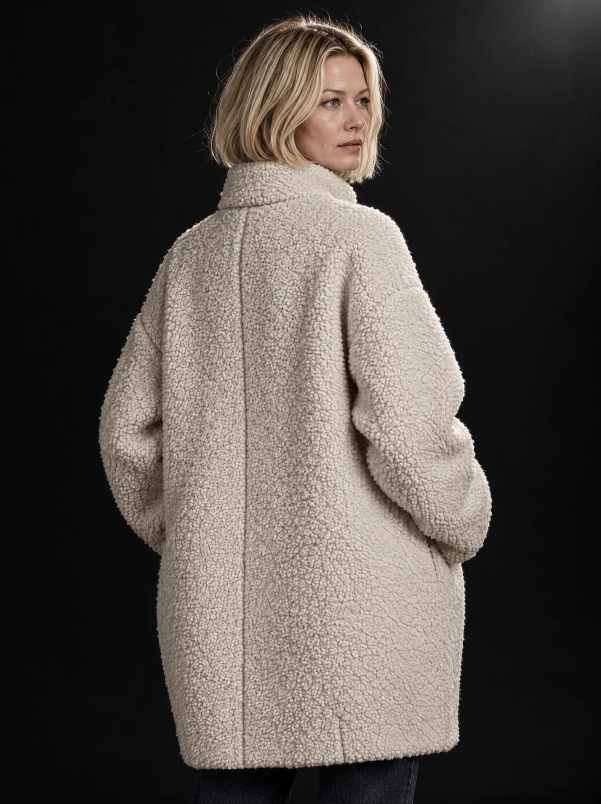 Sophie from behind in the ivory bouclé cocoon coat, looking over her shoulder, in a black studio.