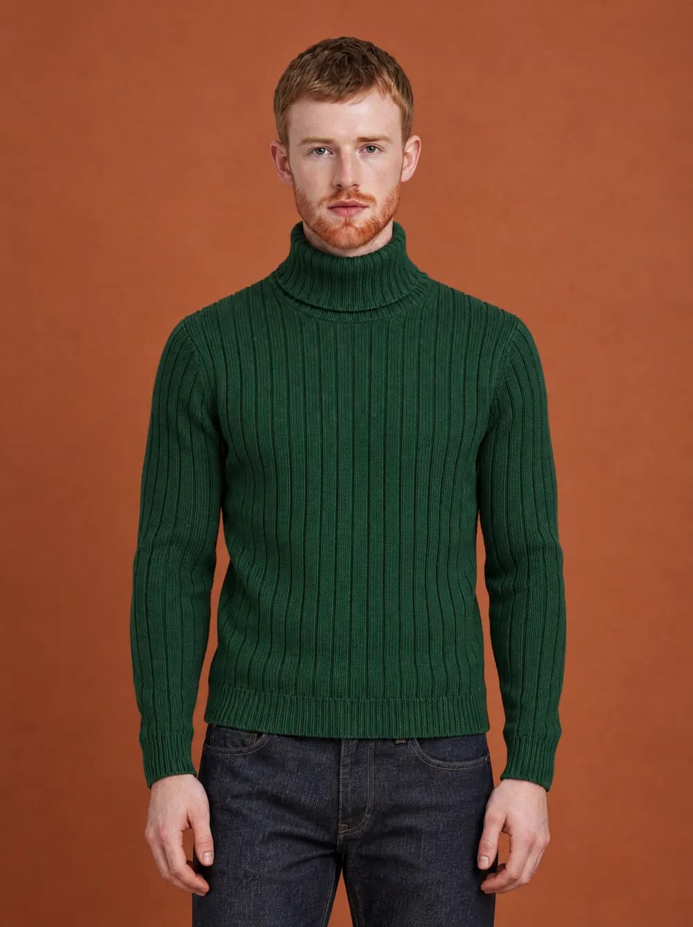 Liam in the green wide-rib turtleneck and the straight dark jeans, against a terracotta studio wall.