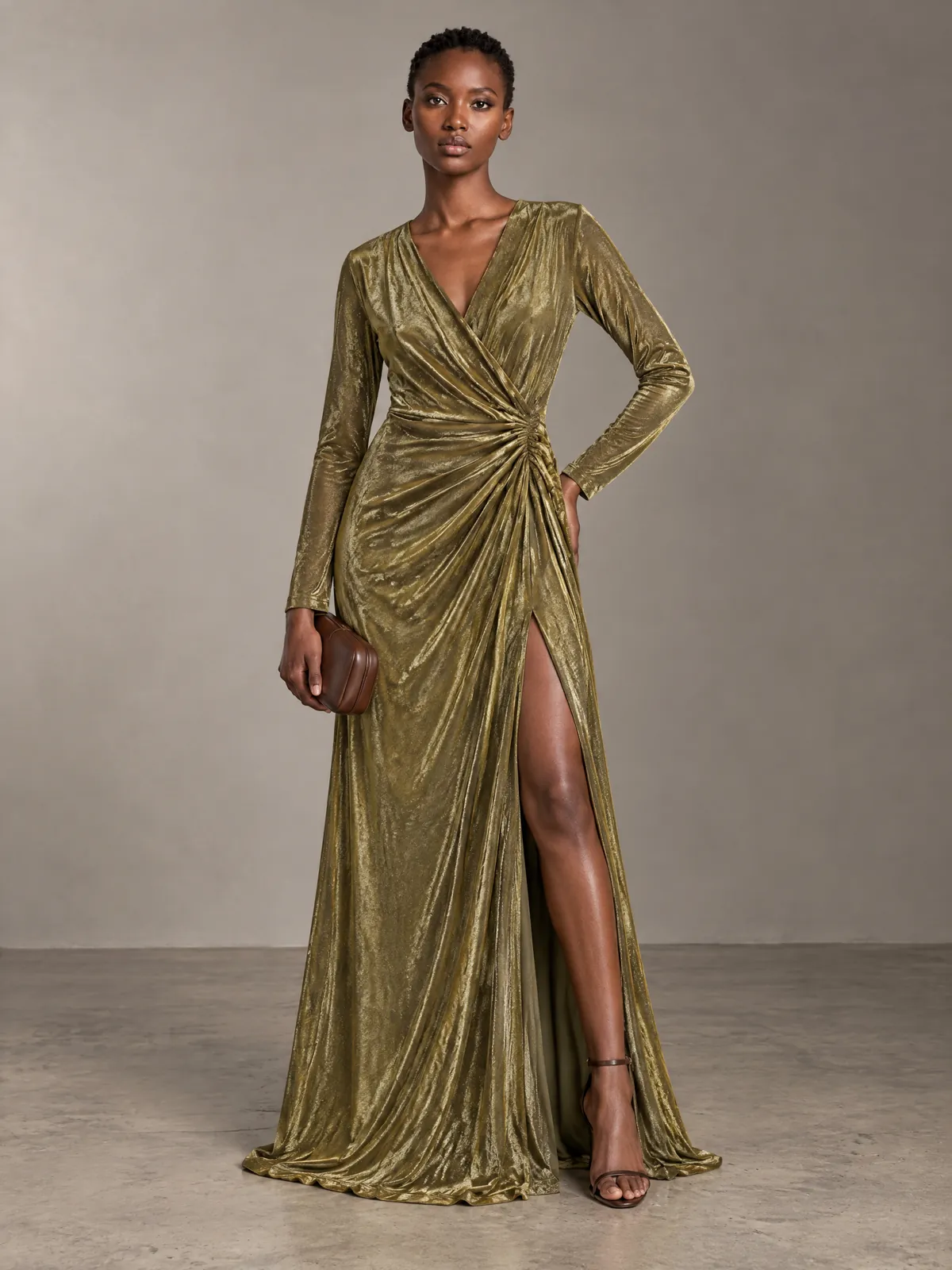 Zola in a long gold jersey wrap gown with a slit, holding a brown clutch, in a grey studio.